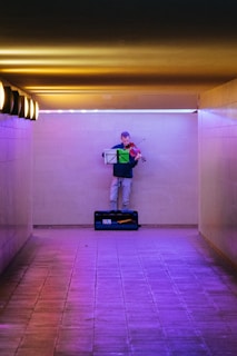 A person stands in an underground passage, playing a violin. They are positioned against a wall, with a music stand in front of them holding sheet music. The corridor is illuminated with warm yellow overhead lights on one side and purple lighting on the floor and lower part of the wall.