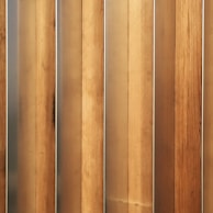 Vertical wooden planks with a natural finish are aligned in a parallel pattern. The wood has visible grain and variation in shades, creating a textured appearance. The planks are evenly spaced and have a warm, earthy tone.