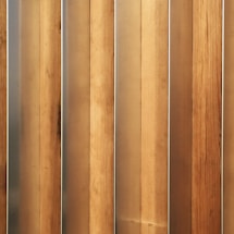 Vertical wooden planks with a natural finish are aligned in a parallel pattern. The wood has visible grain and variation in shades, creating a textured appearance. The planks are evenly spaced and have a warm, earthy tone.