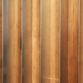 Vertical wooden planks with a natural finish are aligned in a parallel pattern. The wood has visible grain and variation in shades, creating a textured appearance. The planks are evenly spaced and have a warm, earthy tone.