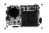 An X-ray view of electronic circuitry with various components visible, including round structures and complex circuit patterns. The image highlights intricate details of a printed circuit board with components like capacitors, resistors, and possibly connectors. The contrast of dark and light areas gives insight into the internal layout.