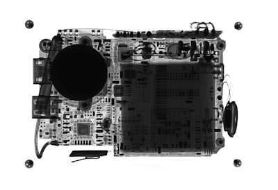 An X-ray view of electronic circuitry with various components visible, including round structures and complex circuit patterns. The image highlights intricate details of a printed circuit board with components like capacitors, resistors, and possibly connectors. The contrast of dark and light areas gives insight into the internal layout.