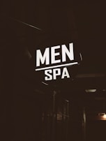 A brightly illuminated sign featuring the words 'MEN SPA' is displayed in a dark, industrial-looking environment. The sign stands out in contrast to the dim surroundings, providing a focus point against the otherwise shadowy backdrop.
