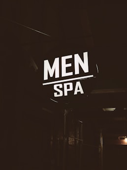 A brightly illuminated sign featuring the words 'MEN SPA' is displayed in a dark, industrial-looking environment. The sign stands out in contrast to the dim surroundings, providing a focus point against the otherwise shadowy backdrop.