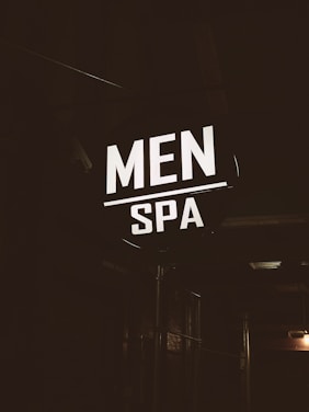 A brightly illuminated sign featuring the words 'MEN SPA' is displayed in a dark, industrial-looking environment. The sign stands out in contrast to the dim surroundings, providing a focus point against the otherwise shadowy backdrop.