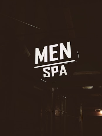 A brightly illuminated sign featuring the words 'MEN SPA' is displayed in a dark, industrial-looking environment. The sign stands out in contrast to the dim surroundings, providing a focus point against the otherwise shadowy backdrop.