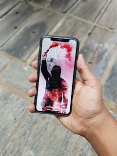 A hand is holding a smartphone displaying a lock screen with a figure standing in a hooded outfit amidst red smoke. The background includes clear tiles on the ground.