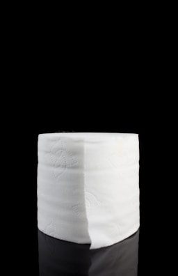 A single roll of white toilet paper centered against a black background, slightly reflecting the surface it's placed on.
