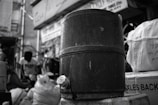 A large, cylindrical metal container with a valve at the bottom sits prominently in the foreground. In the background, blurred figures and bags are visible, hinting at a busy urban or market setting. The entire scene is in black and white, emphasizing textures and contrasts.