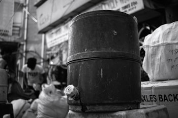 A large, cylindrical metal container with a valve at the bottom sits prominently in the foreground. In the background, blurred figures and bags are visible, hinting at a busy urban or market setting. The entire scene is in black and white, emphasizing textures and contrasts.