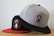 Two baseball caps are positioned on a light-colored surface. The cap in the foreground is gray with a black visor and features a circular logo with a bearded man's face. The cap in the background is black with a red visor and also has a similar circular logo of a bearded man.