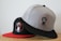 Two baseball caps are positioned on a light-colored surface. The cap in the foreground is gray with a black visor and features a circular logo with a bearded man's face. The cap in the background is black with a red visor and also has a similar circular logo of a bearded man.