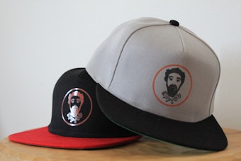 Two baseball caps are positioned on a light-colored surface. The cap in the foreground is gray with a black visor and features a circular logo with a bearded man's face. The cap in the background is black with a red visor and also has a similar circular logo of a bearded man.