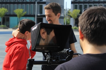 Three individuals are engaged in a video shoot outdoors, with two looking at a monitor. The monitor displays a close-up of an ear. One person is wearing a red hoodie and receiving direction or comfort from another. The background includes modern buildings and trees.