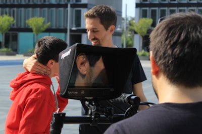 Three individuals are engaged in a video shoot outdoors, with two looking at a monitor. The monitor displays a close-up of an ear. One person is wearing a red hoodie and receiving direction or comfort from another. The background includes modern buildings and trees.