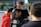 Three individuals are engaged in a video shoot outdoors, with two looking at a monitor. The monitor displays a close-up of an ear. One person is wearing a red hoodie and receiving direction or comfort from another. The background includes modern buildings and trees.