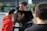 Three individuals are engaged in a video shoot outdoors, with two looking at a monitor. The monitor displays a close-up of an ear. One person is wearing a red hoodie and receiving direction or comfort from another. The background includes modern buildings and trees.