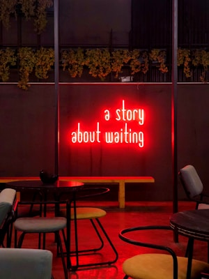 Neon red text on a dark wall reads 'a story about waiting'. The setting appears to be an indoor cafe or bar with empty tables and chairs. Greenery is seen hanging above the neon sign, creating a cozy and intimate atmosphere.