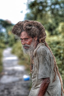 An elderly man with a long, gray beard and thick, matted hair stands wearing a worn, light-colored shirt. His expression is contemplative, and he appears to be outdoors on a blurred pathway, surrounded by green foliage.