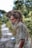 An elderly man with a long, gray beard and thick, matted hair stands wearing a worn, light-colored shirt. His expression is contemplative, and he appears to be outdoors on a blurred pathway, surrounded by green foliage.