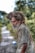 An elderly man with a long, gray beard and thick, matted hair stands wearing a worn, light-colored shirt. His expression is contemplative, and he appears to be outdoors on a blurred pathway, surrounded by green foliage.