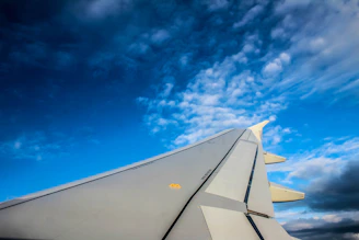 A dynamic shot of an airplane wing cutting through a bright blue sky with fluffy clouds.