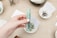 A hand holding a polished green crystal obelisk, surrounded by small bowls containing stones and cards with text. Some items in the background are slightly out of focus, enhancing the focus on the crystal.