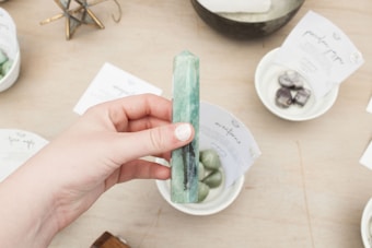 A hand holding a polished green crystal obelisk, surrounded by small bowls containing stones and cards with text. Some items in the background are slightly out of focus, enhancing the focus on the crystal.