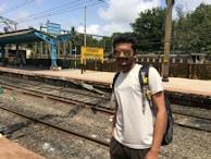 A person wearing sunglasses and a backpack stands near a railway platform. The platform has blue and yellow signs displaying the name 'Lonavla' in multiple languages. The tracks are visible in the foreground and trees are seen in the background under a partly cloudy sky.