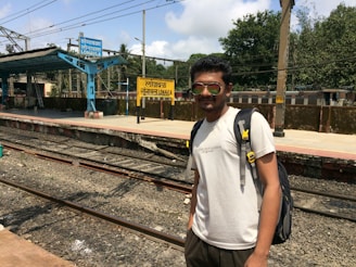 A person wearing sunglasses and a backpack stands near a railway platform. The platform has blue and yellow signs displaying the name 'Lonavla' in multiple languages. The tracks are visible in the foreground and trees are seen in the background under a partly cloudy sky.