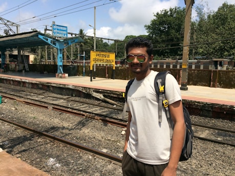 A person wearing sunglasses and a backpack stands near a railway platform. The platform has blue and yellow signs displaying the name 'Lonavla' in multiple languages. The tracks are visible in the foreground and trees are seen in the background under a partly cloudy sky.