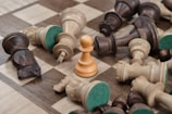 A wooden chessboard is scattered with fallen chess pieces, both dark and light, surrounding a single upright white pawn. The board's squares are alternating shades of brown and beige, and the felt bottoms of the pieces are visible.