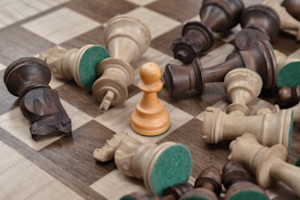 A wooden chessboard is scattered with fallen chess pieces, both dark and light, surrounding a single upright white pawn. The board's squares are alternating shades of brown and beige, and the felt bottoms of the pieces are visible.