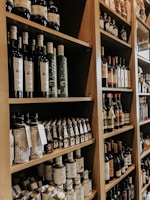 Shelves filled with an assortment of bottles, including olive oil, wine, and various other condiments, neatly arranged in a wooden shelving unit. The bottles vary in size and design, many with intricate labels. The setting appears to be a specialty food store or a gourmet market.