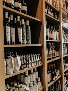 Shelves filled with an assortment of bottles, including olive oil, wine, and various other condiments, neatly arranged in a wooden shelving unit. The bottles vary in size and design, many with intricate labels. The setting appears to be a specialty food store or a gourmet market.