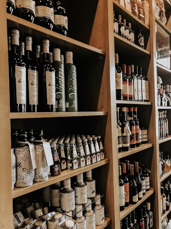 Shelves filled with an assortment of bottles, including olive oil, wine, and various other condiments, neatly arranged in a wooden shelving unit. The bottles vary in size and design, many with intricate labels. The setting appears to be a specialty food store or a gourmet market.