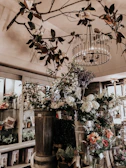 An interior space filled with an assortment of potted plants and flowers. The ceiling is adorned with branches and white flowers, complemented by a large, ornate chandelier. Various greenery and floral arrangements are displayed on shelves, and large vases hold vibrant blooms. The space has a cozy and rustic atmosphere.