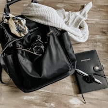 A black backpack lies on a wooden surface with a camera partially visible inside. A patterned scarf is draped on top of the bag, and a knitted sweater is next to it. To the right, a black notebook with 'NOTEBOOK' written on the cover is accompanied by a pen with a cap attached by a string.