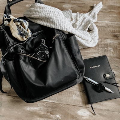 A black backpack lies on a wooden surface with a camera partially visible inside. A patterned scarf is draped on top of the bag, and a knitted sweater is next to it. To the right, a black notebook with 'NOTEBOOK' written on the cover is accompanied by a pen with a cap attached by a string.