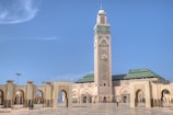 A grand mosque with a tall minaret adorned with intricate designs stands under a clear blue sky. The architectural style is traditional Islamic, featuring detailed patterns and a combination of sandstone and green-tiled roofs. The surrounding area is an open courtyard with a few people visible, emphasizing the scale and majesty of the structure.