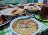 A table is set with various plates of food, including a steaming bowl of noodle soup with meat and garnishes, fried items like tofu and tempeh, and a glass of iced tea. The background features a checker-patterned tablecloth and a colorful assortment of dishes.