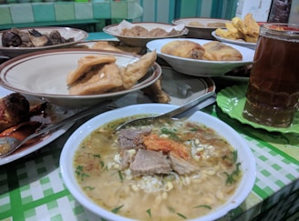 A table is set with various plates of food, including a steaming bowl of noodle soup with meat and garnishes, fried items like tofu and tempeh, and a glass of iced tea. The background features a checker-patterned tablecloth and a colorful assortment of dishes.
