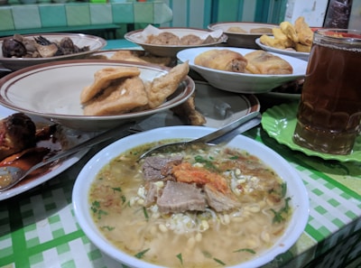 A table is set with various plates of food, including a steaming bowl of noodle soup with meat and garnishes, fried items like tofu and tempeh, and a glass of iced tea. The background features a checker-patterned tablecloth and a colorful assortment of dishes.