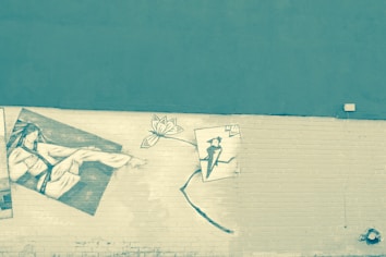 A mural on a brick wall features sketches. On the left side, a reclining figure is depicted. In the center, there is a stylized flower, and on the right side, a smaller figure appears to be holding an umbrella or a similar object.