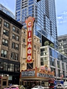 A classic theater marquee in downtown, prominently displaying the name 'Chicago' in bold, vintage-style red lettering. The theater is surrounded by tall, modern skyscrapers with reflective glass windows. The marquee features upcoming performances, including ones by Hasan Minhaj and Lucinda Williams. Bright streetlights and a bustling urban environment are visible in the scene.