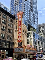 A classic theater marquee in downtown, prominently displaying the name 'Chicago' in bold, vintage-style red lettering. The theater is surrounded by tall, modern skyscrapers with reflective glass windows. The marquee features upcoming performances, including ones by Hasan Minhaj and Lucinda Williams. Bright streetlights and a bustling urban environment are visible in the scene.