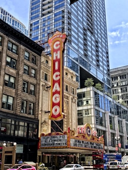 A classic theater marquee in downtown, prominently displaying the name 'Chicago' in bold, vintage-style red lettering. The theater is surrounded by tall, modern skyscrapers with reflective glass windows. The marquee features upcoming performances, including ones by Hasan Minhaj and Lucinda Williams. Bright streetlights and a bustling urban environment are visible in the scene.