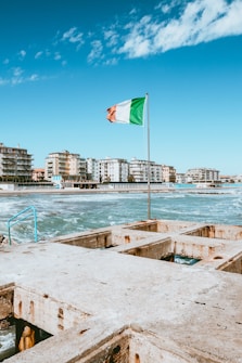 A coastal scene featuring a concrete pier with a hole-filled surface extending into the water. An Italian flag flutters on a pole against a bright blue sky. In the background, a row of modern apartment buildings lines the waterfront, and waves crash against the shore.
