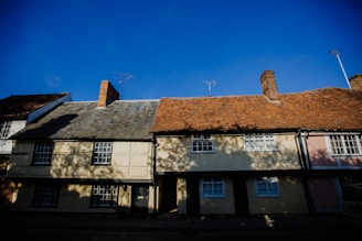 A row of traditional English cottages, each with distinct sloped roofs and chimneys, depicted under a clear blue sky. Shadows from trees fall across the cream-colored walls, and TV antennas are visible atop the rooftops.