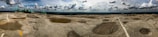 A vast rocky terrain stretches across the image with distinctive craters and a few painted markings. The sky above is partly cloudy, casting dynamic shadows on the ground. Structures with teal roofs are visible on the left side, likely facilities or observation points.